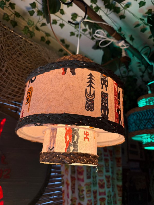 May include: A pendant lamp with a two-tiered lampshade featuring a light tan fabric adorned with black and red tiki-style figures. The top and bottom edges are trimmed with a dark woven material. The lamp hangs from a black hook.